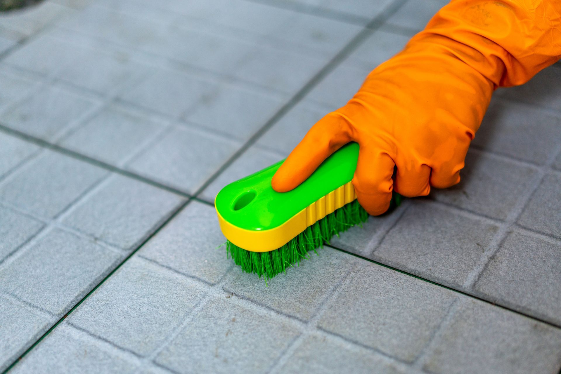 A person wearing orange gloves is cleaning a tile floor with a green and yellow brush.