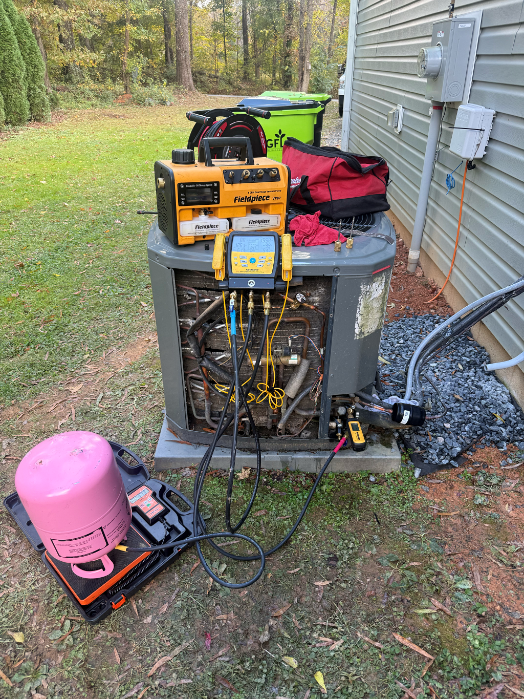 HVAC technician working on an AC unit outdoors. Tools, gauges, and a pink tank are visible.