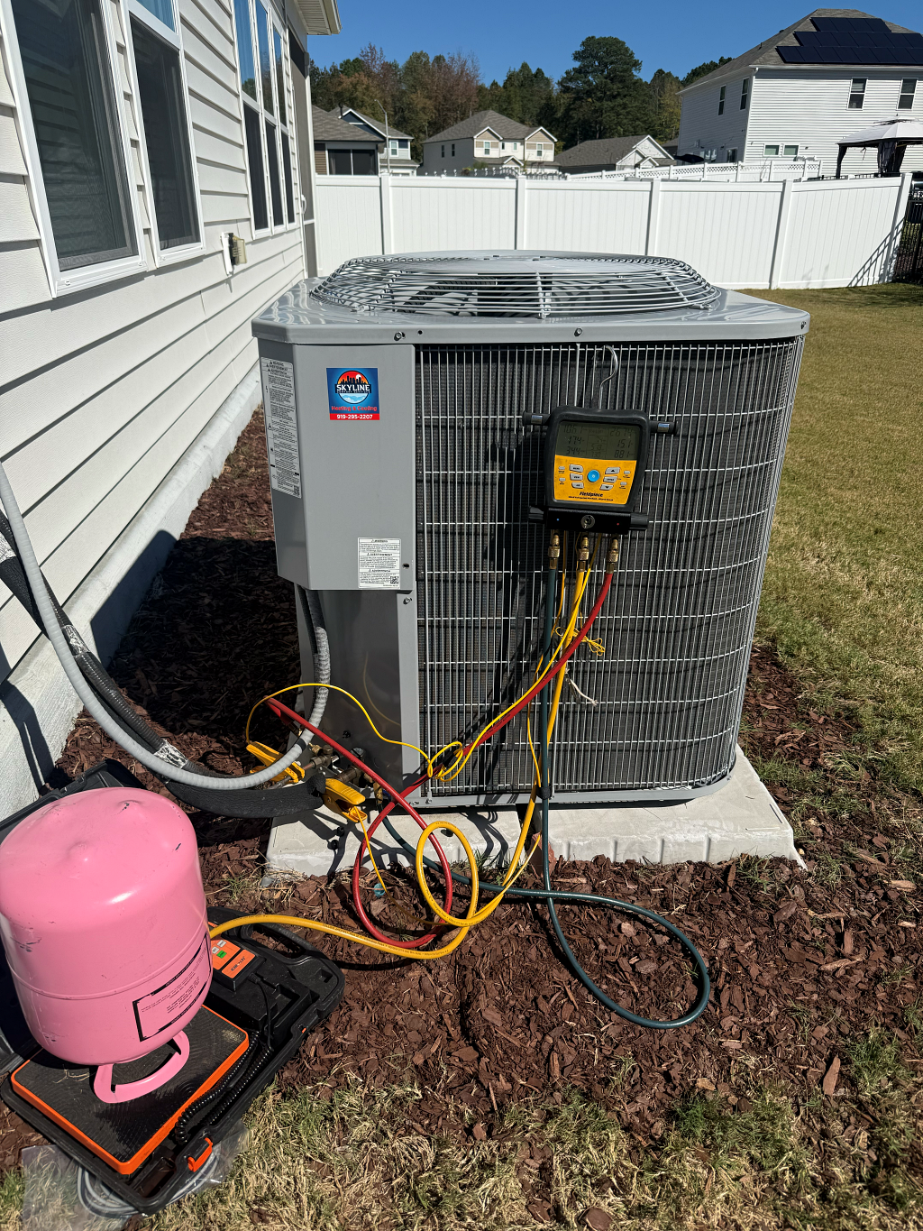 An HVAC unit with gauges attached, next to a pink refrigerant tank, in a yard with a white fence.