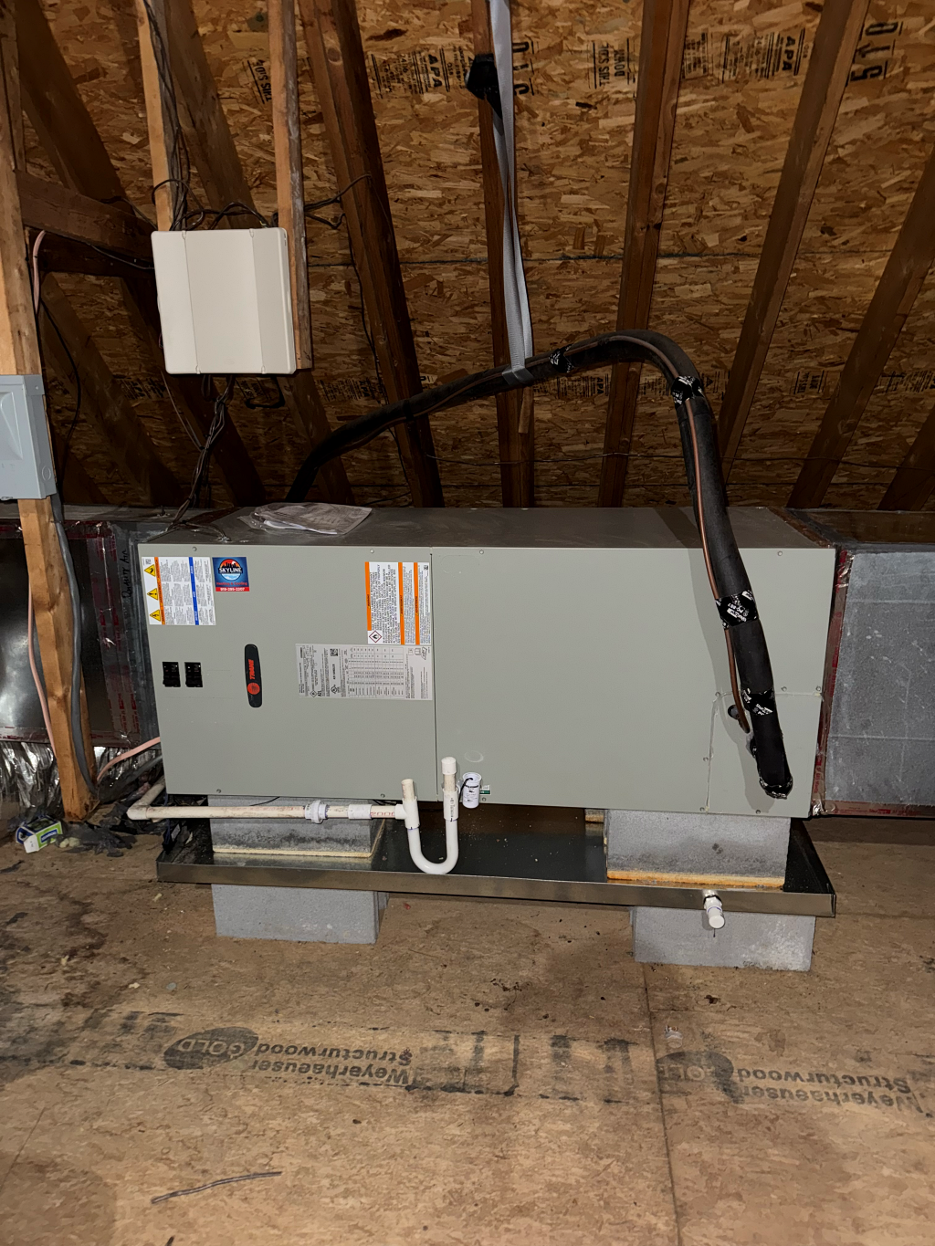 An HVAC system in an attic. Gray unit on concrete blocks, with black ductwork and white drain pipes.