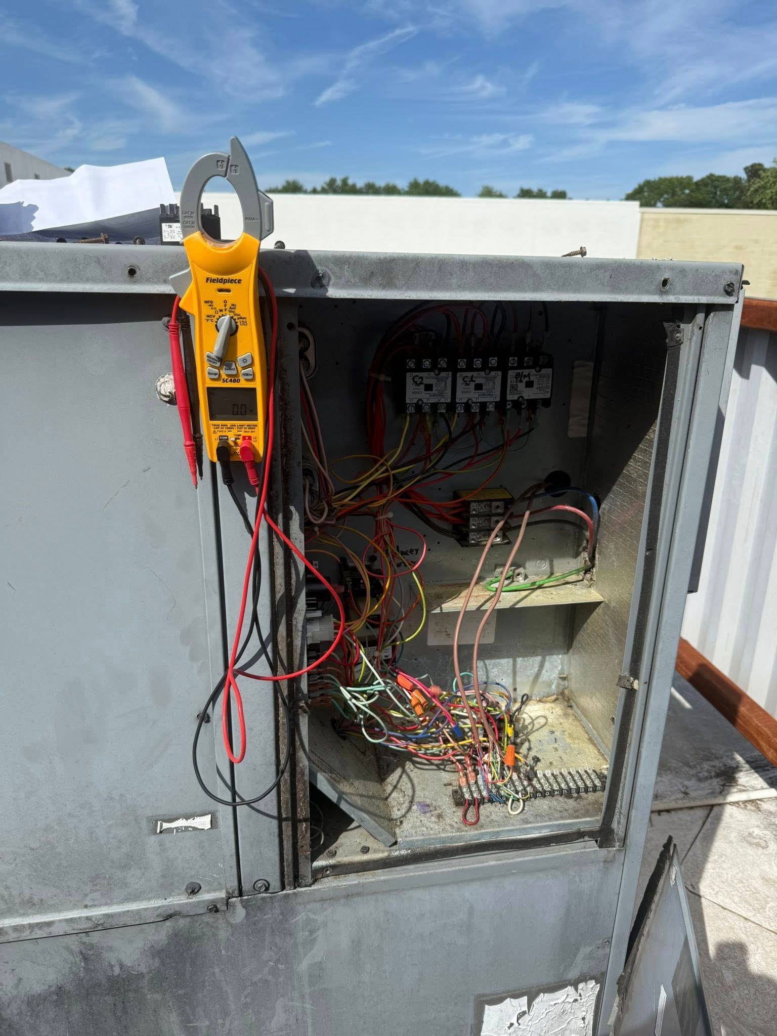 Skyline Comfort Services LLC - HVAC electrical panel open, with a yellow multimeter clamped around a wire on a rooftop.