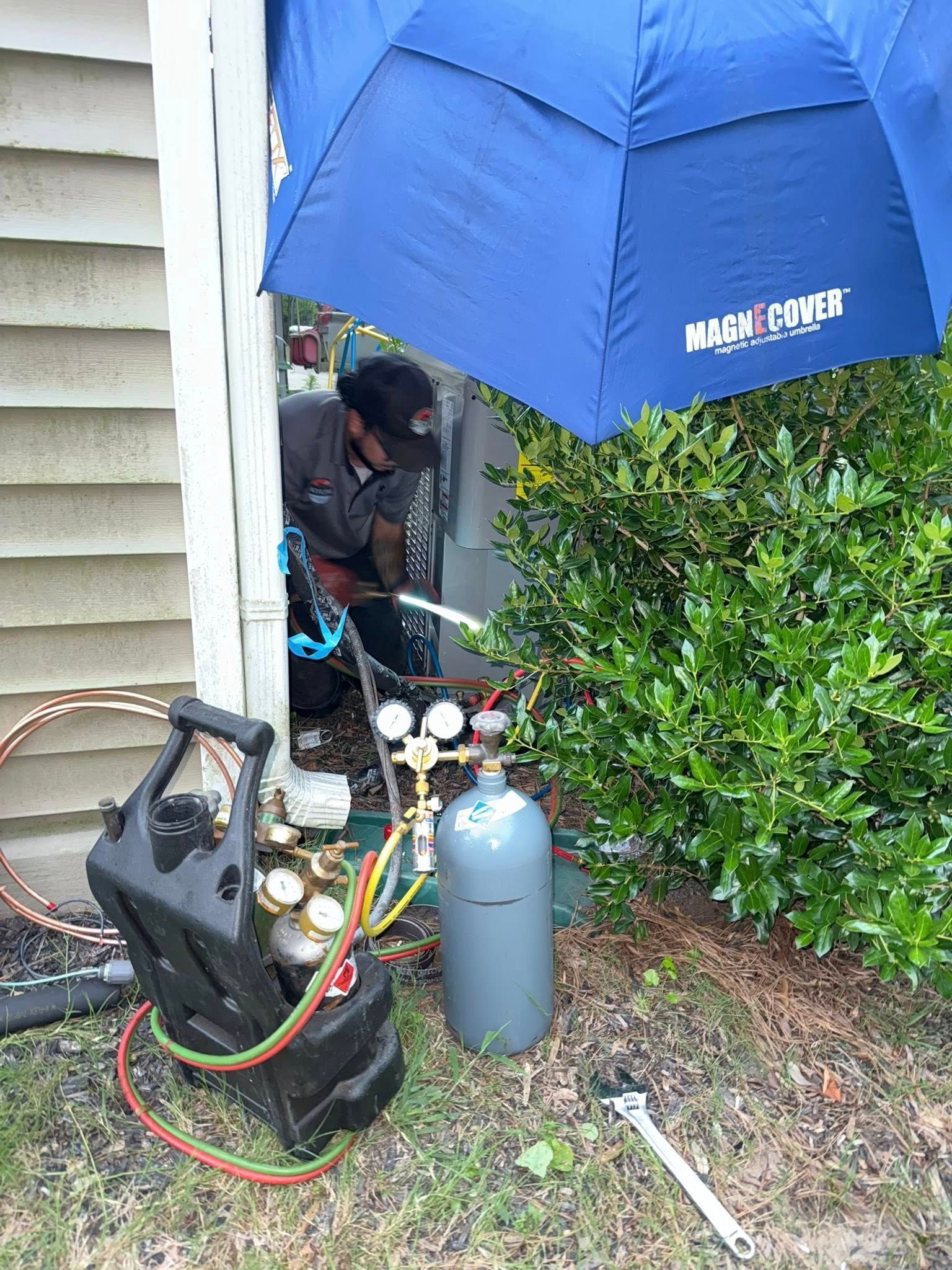 Skyline Comfort Services LLC - HVAC technician using a torch, brazing copper pipes, under a blue umbrella near a green bush.