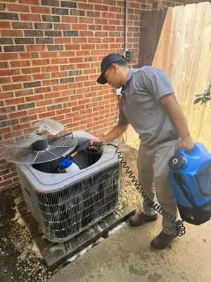 Skyline Comfort Services LLC - Man cleaning an AC unit with a hose, next to a brick wall and wooden fence.