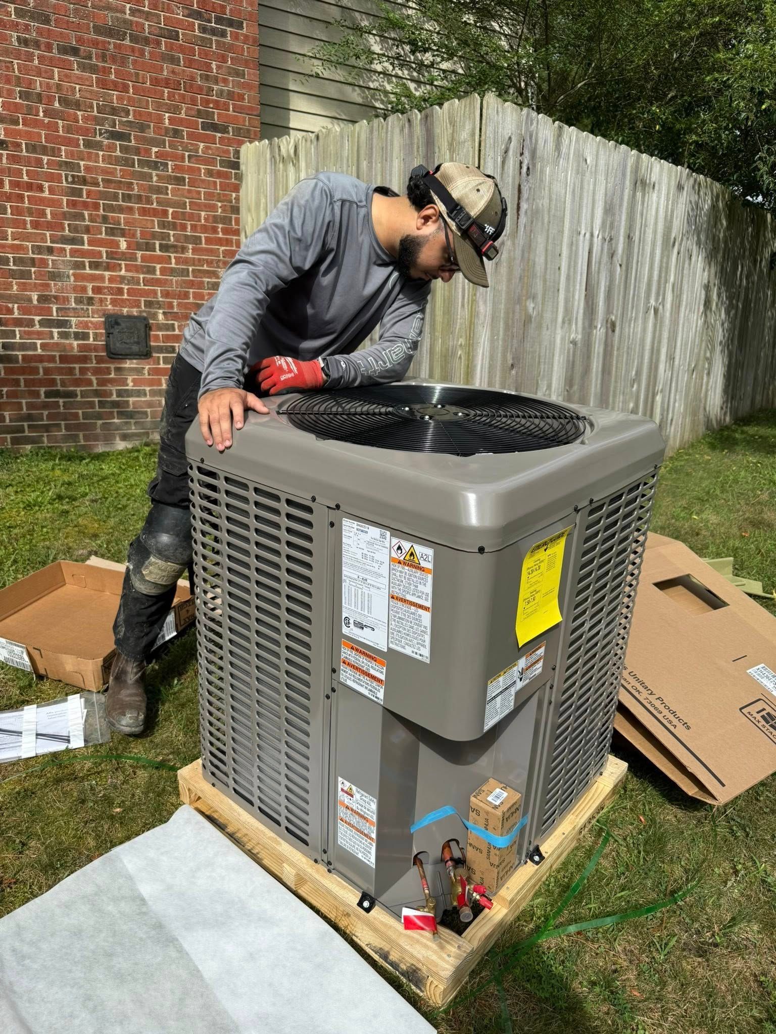 Skyline Comfort Services LLC - HVAC technician inspecting an air conditioner in a backyard. Wears a hat, kneepads and gloves.