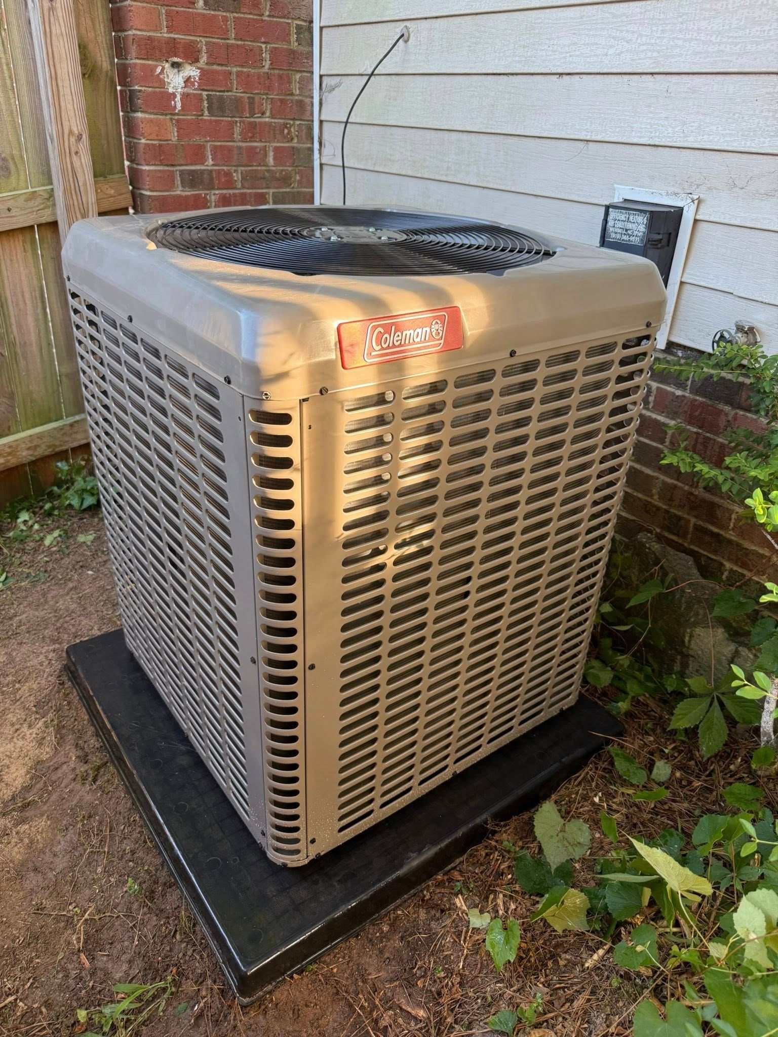 Skyline Comfort Services LLC - Tan air conditioning unit outside on a black pad next to a building and some greenery.