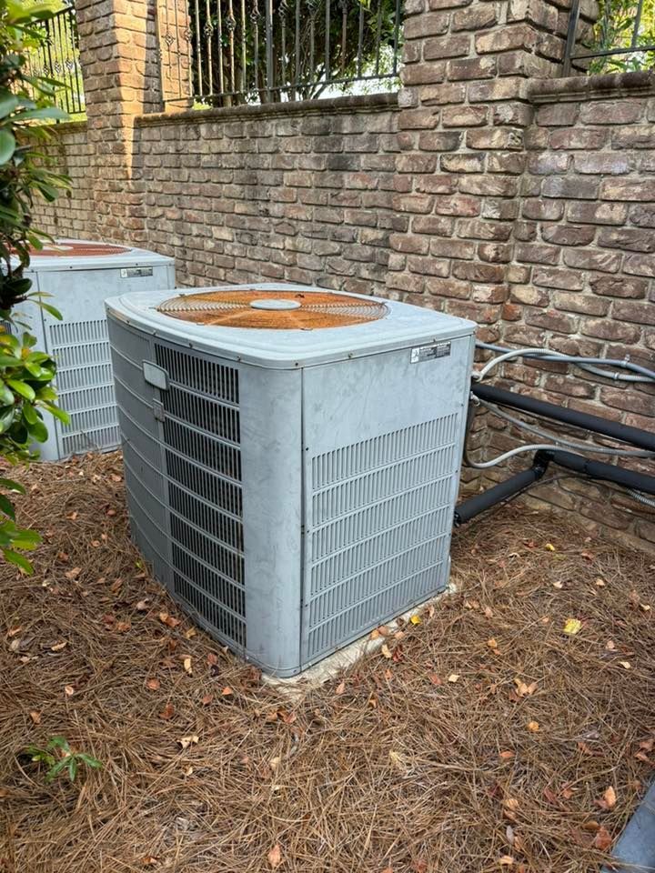 Skyline Comfort Services LLC - Two gray air conditioning units sitting outside on pine straw, near a brick wall.