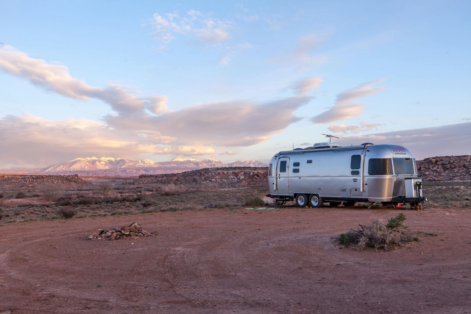 Silver Airstream trailer parked on red dirt, desert landscape with mountains under a cloudy sky.