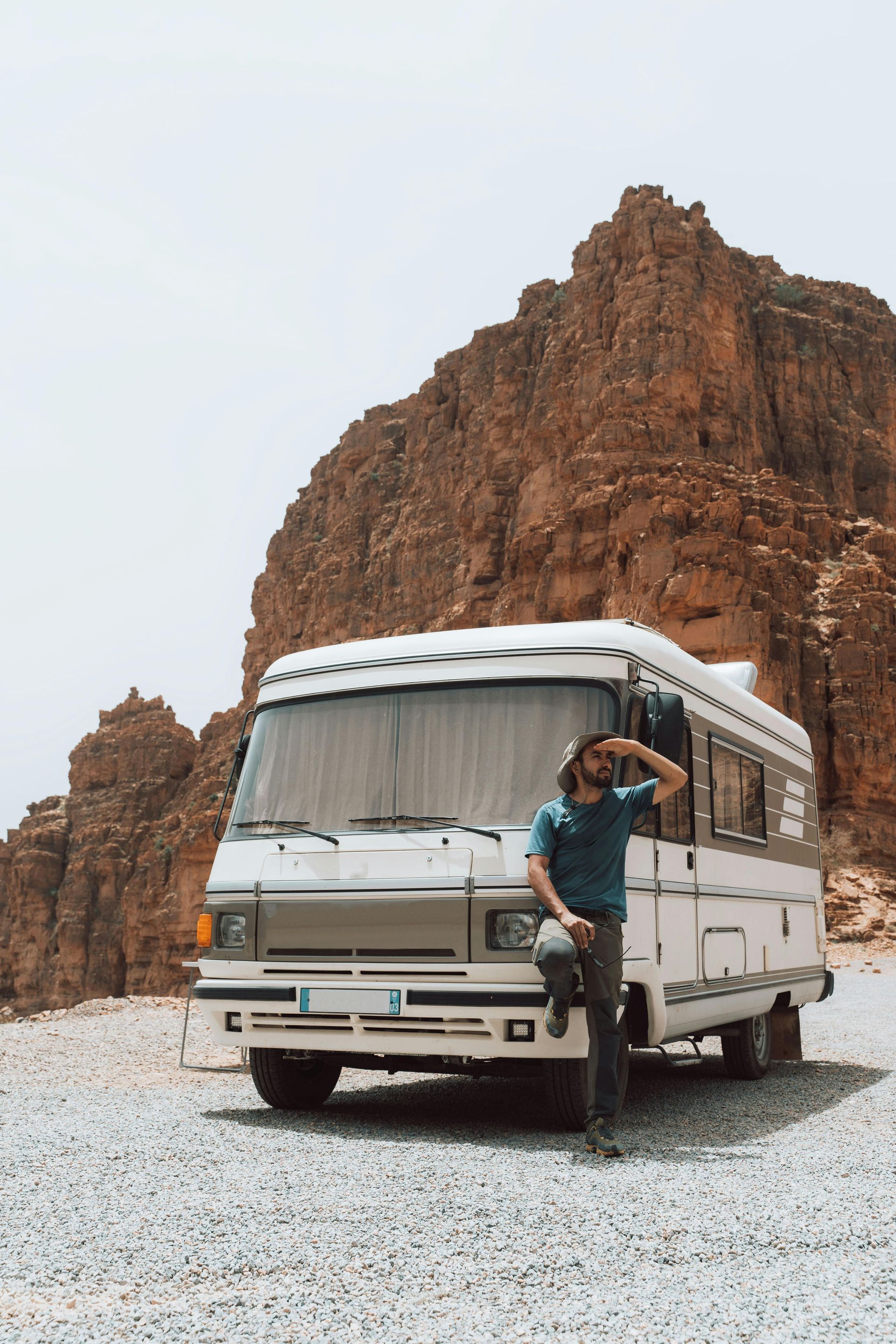 Man in blue shirt, shades eyes, leans on a white RV parked in a rocky desert landscape near a large rock formation.