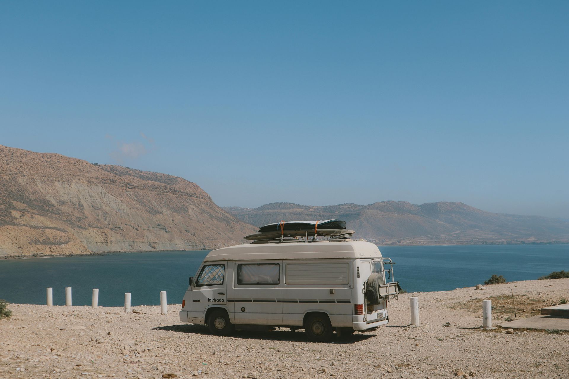 White camper van parked near a body of water with mountains under a bright blue sky.