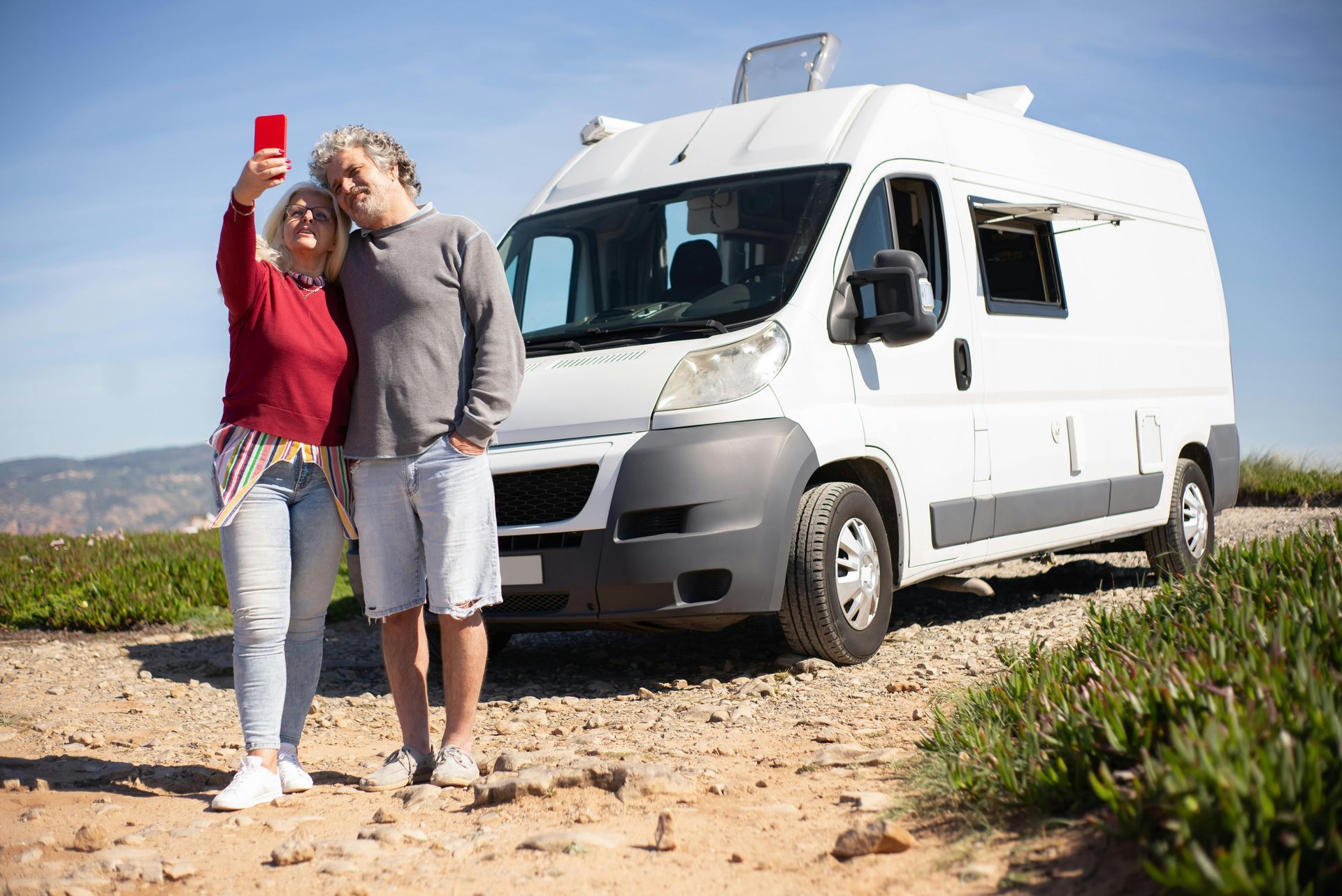 Couple taking a selfie in front of a white camper van on a sunny day.