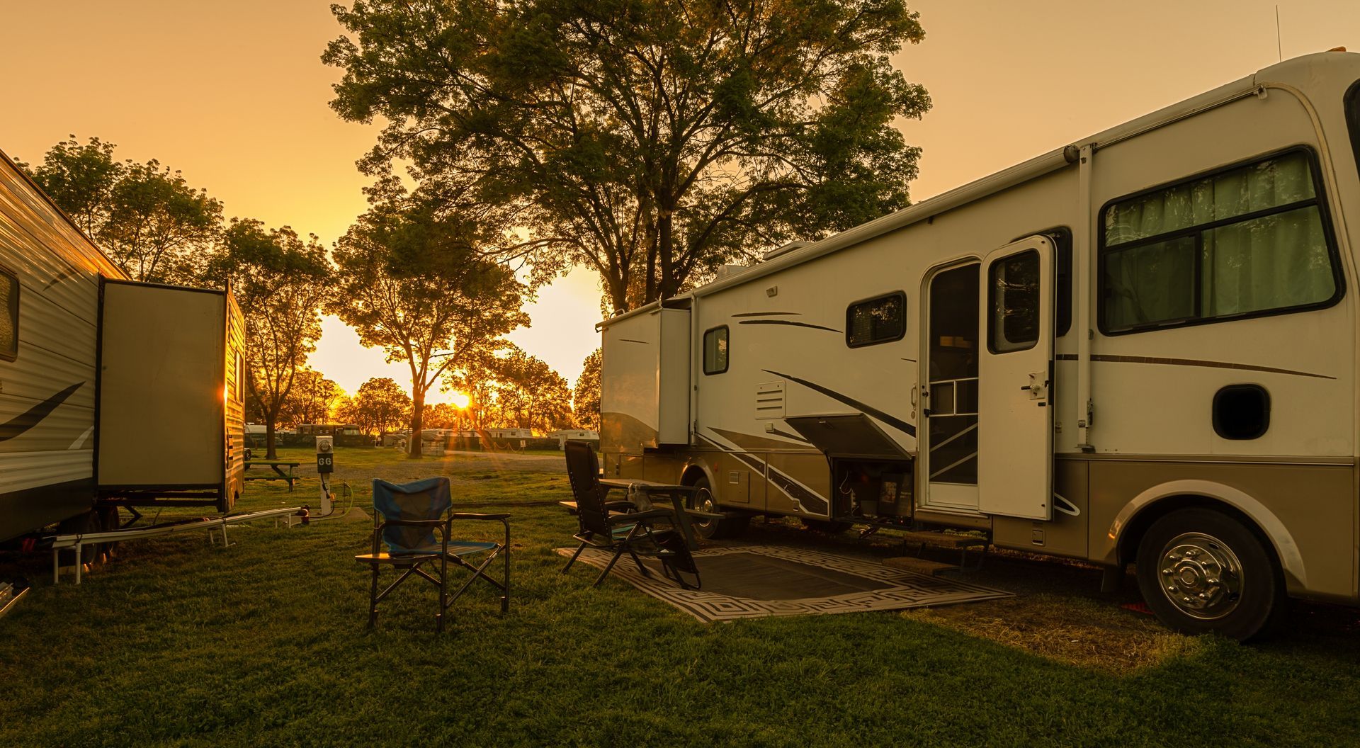 Campground at sunset, featuring recreational vehicles parked on grass, folding chairs, and a tree.