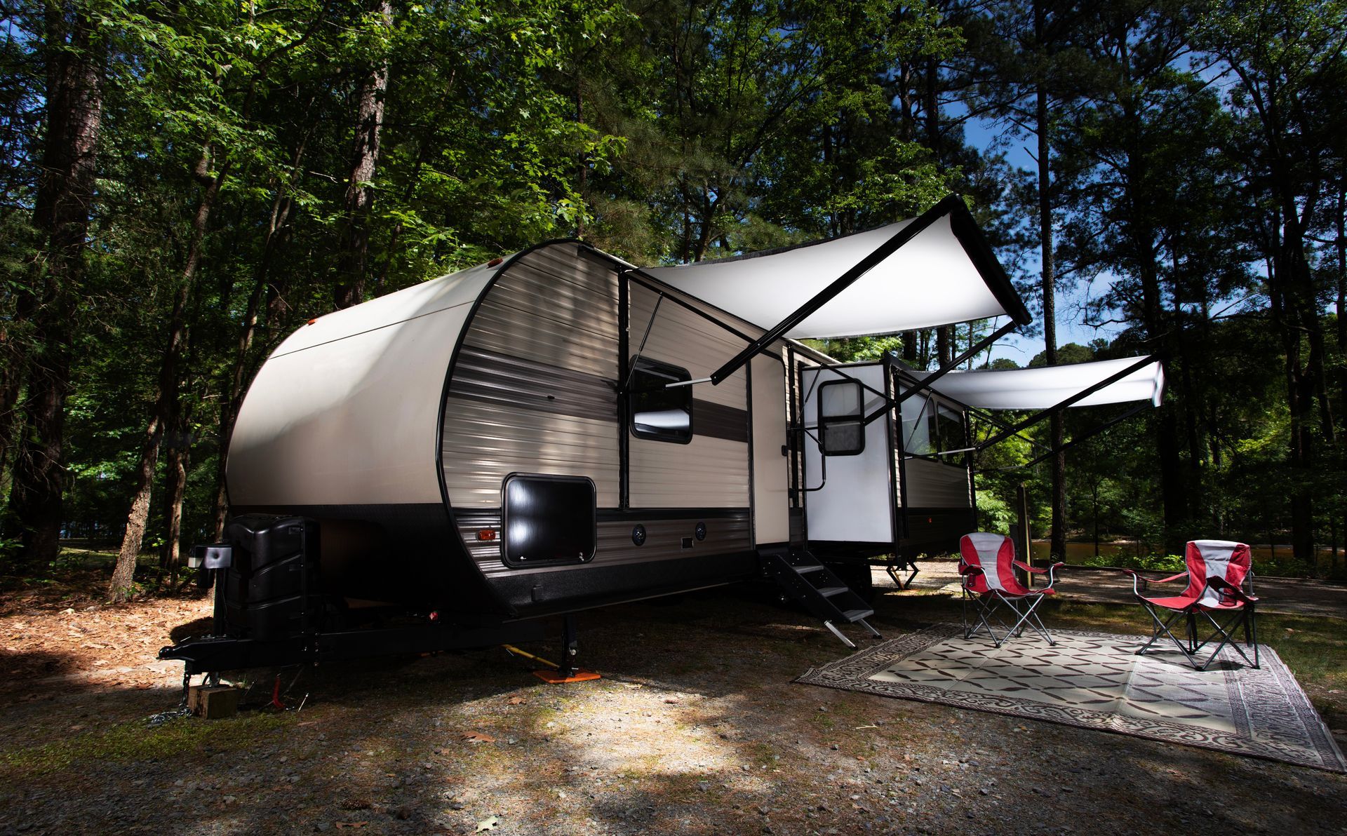 RV camper with awning extended in a wooded campground setting; two camp chairs are set out.
