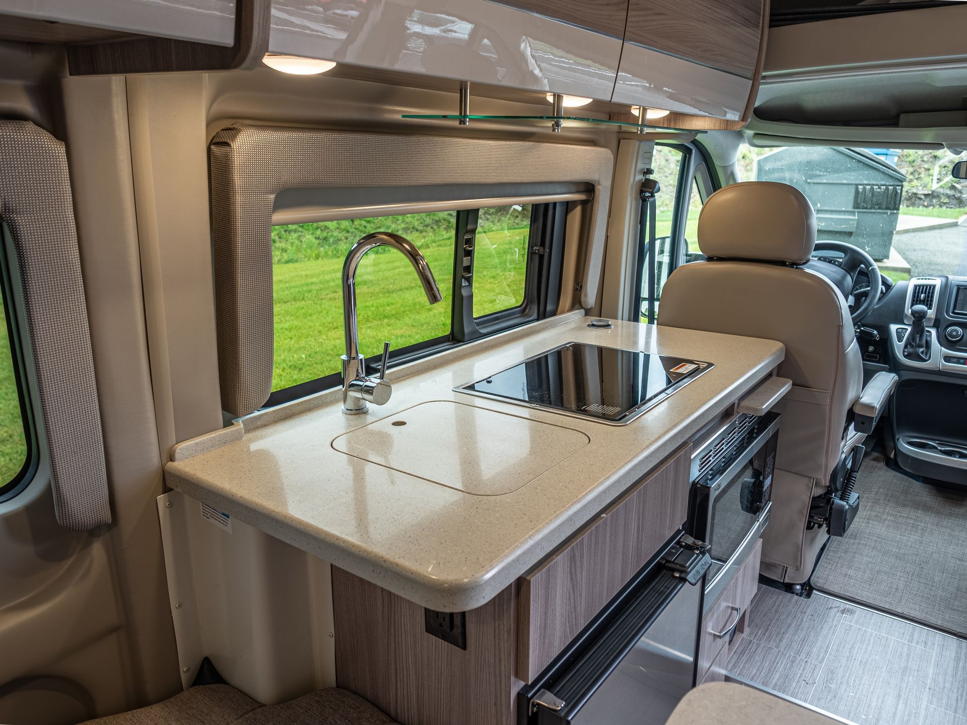 RV interior with a kitchen counter, sink, stove, window, and driver's seat.