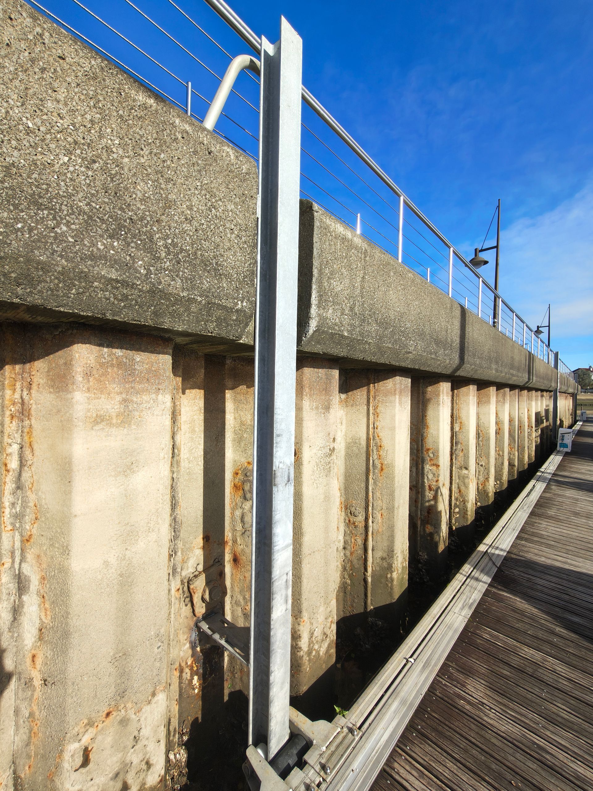 A tall metal rail, with a gray concrete wall and rusty supports. Blue sky and wood planks complete the scene.