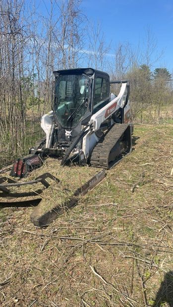 A compact track loader with brush-clearing attachment working in a grassy field under a blue sky