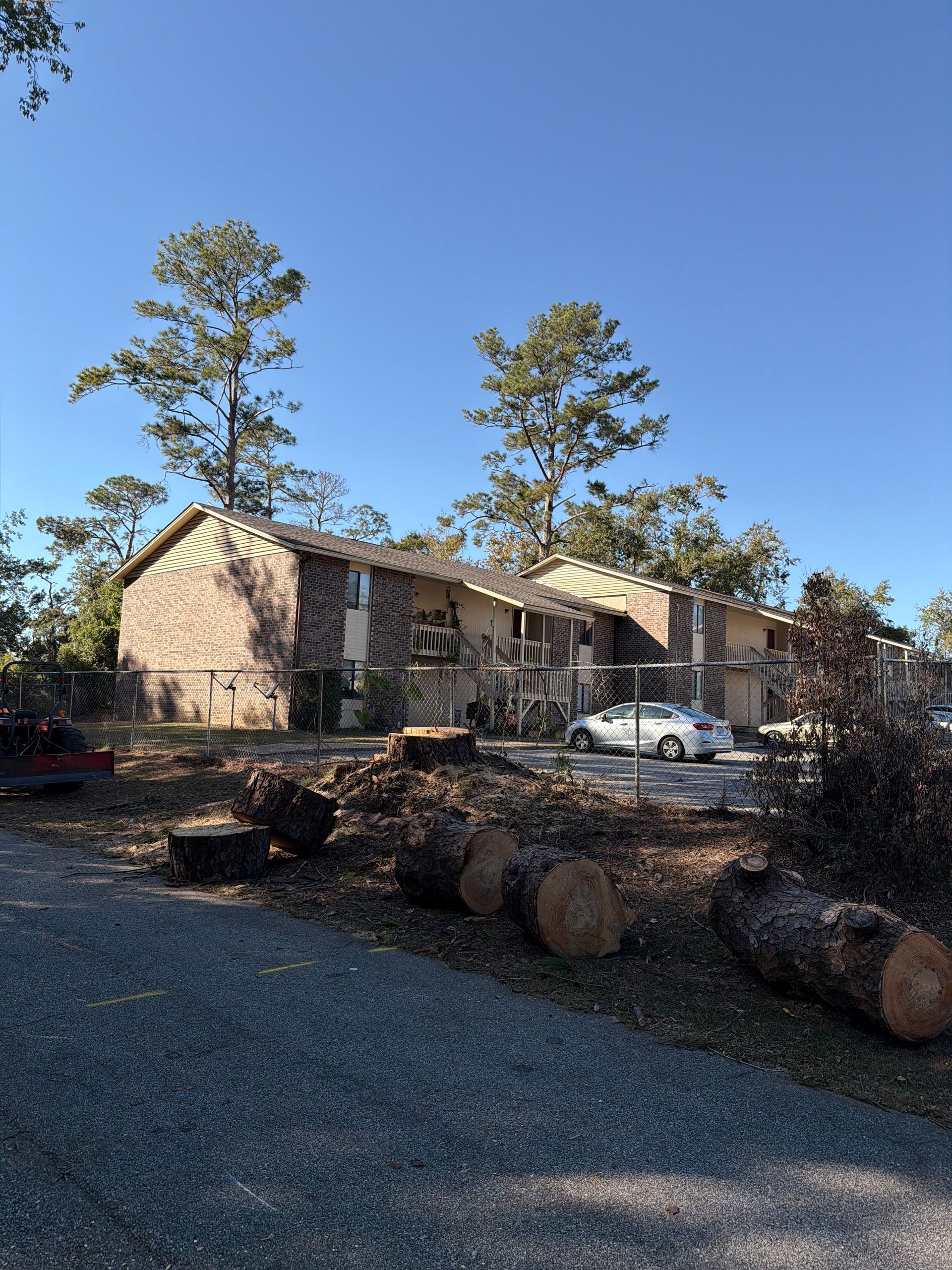 Apartment building with cut tree logs in the foreground, parked car, clear blue sky.