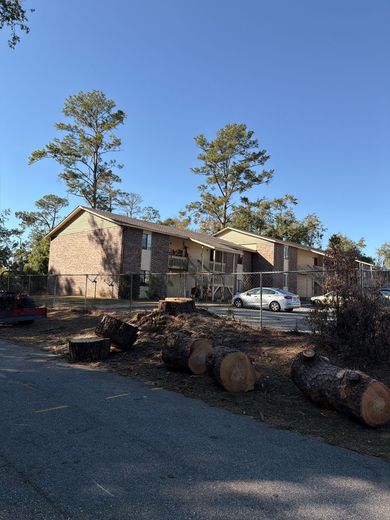 Apartment building with cut tree logs in the foreground, parked car, clear blue sky.