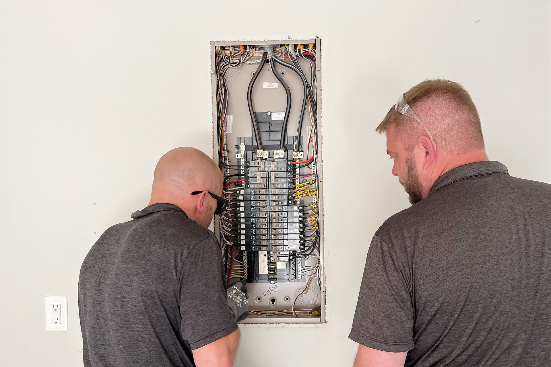 Two men are working on an electrical panel on a wall.