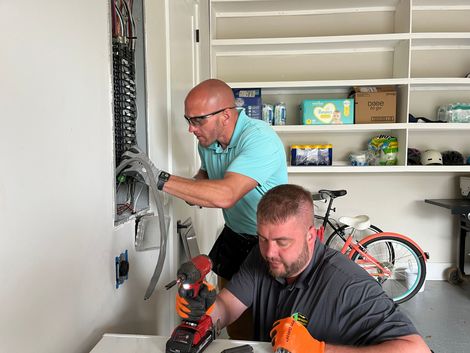 Two men are working on a electrical panel in a garage.