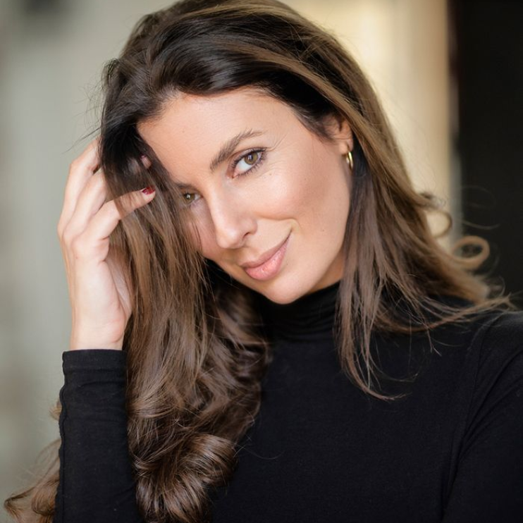 Woman with brown hair in a black turtleneck touches her hair, smiling.