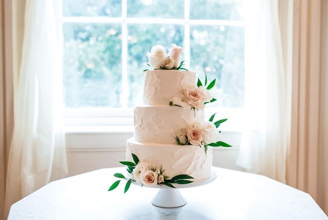 A wedding cake is sitting on a white table in front of a window.