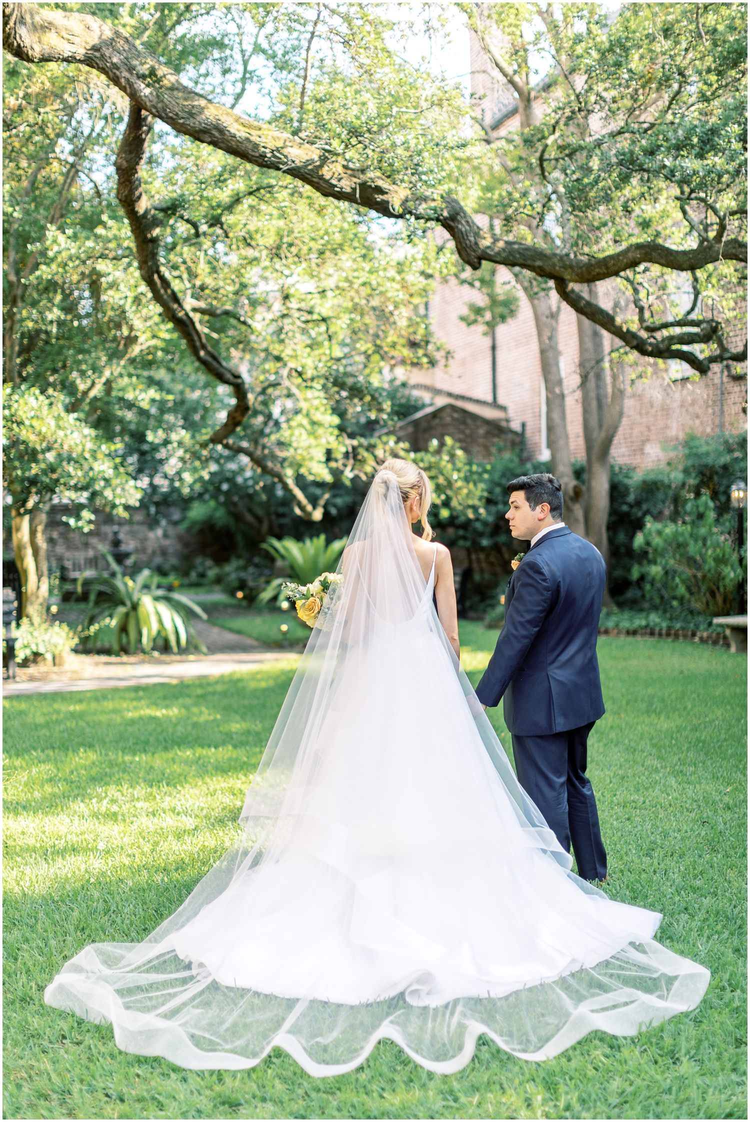 A bride and groom are standing in the grass under a tree . the bride is wearing a long veil.