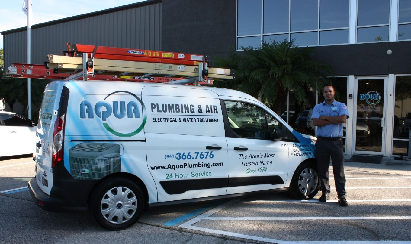 Man stands by a white AQUA Plumbing & Air van with ladder on roof; building in the background.