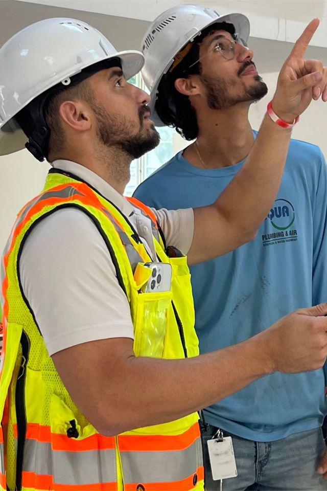 Two construction workers in hard hats examine a ceiling. One points up, wearing a safety vest.