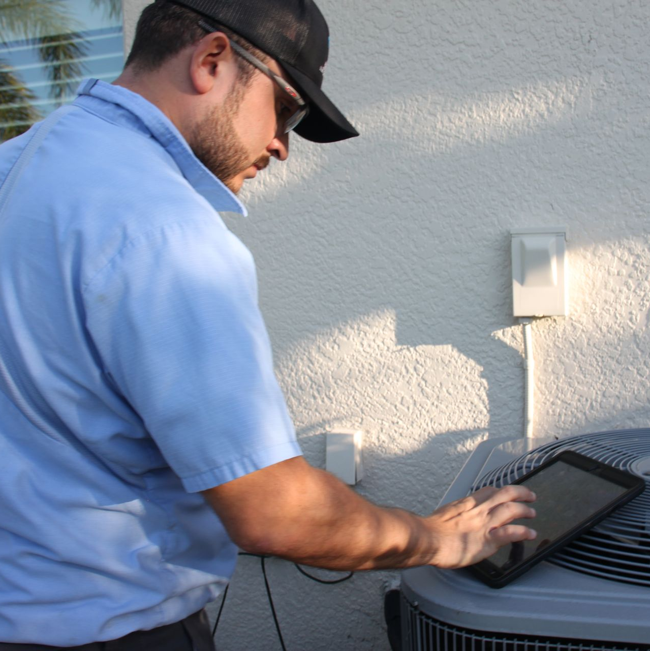 HVAC technician in blue shirt and cap uses a tablet to inspect an air conditioning unit outside a building.