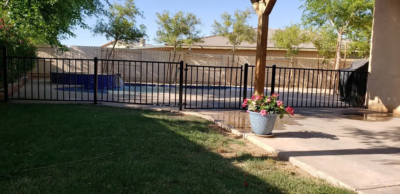 Backyard with a pool, black fence, green lawn, and potted flowers on a patio.
