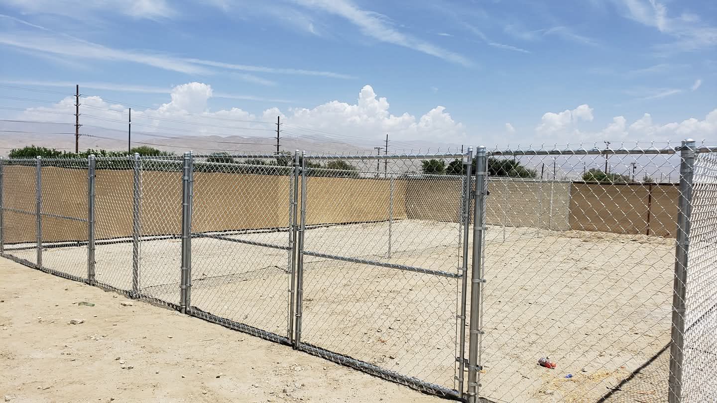 Chain-link fence with beige privacy screening encloses an empty dirt lot under a blue sky.