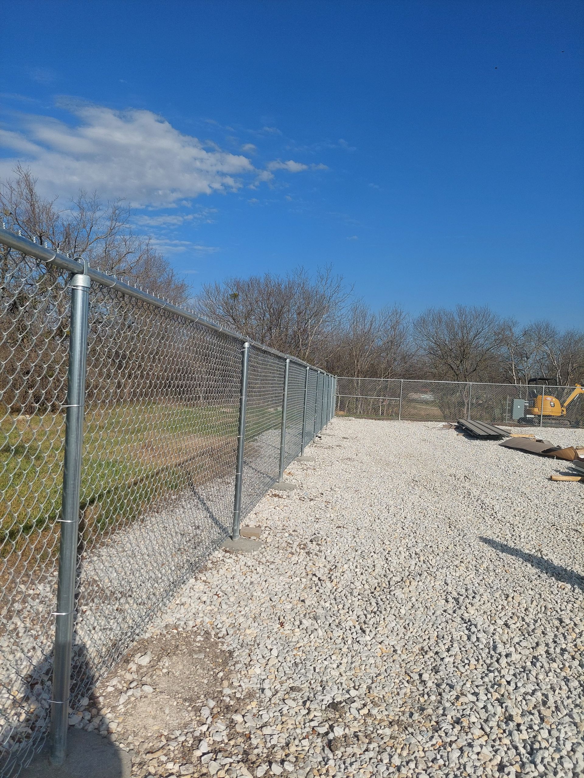 A chain-link fence borders a gravel path under a blue sky. Bare trees and construction materials are visible in the distance.