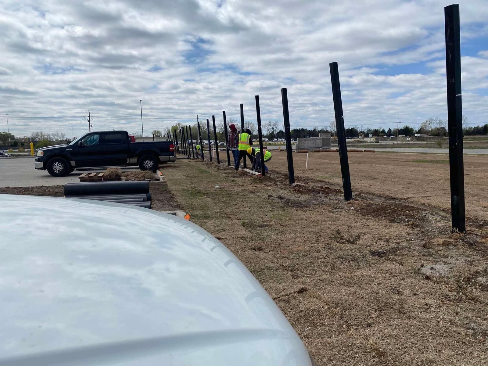 Workers installing black fence posts next to a parking lot.