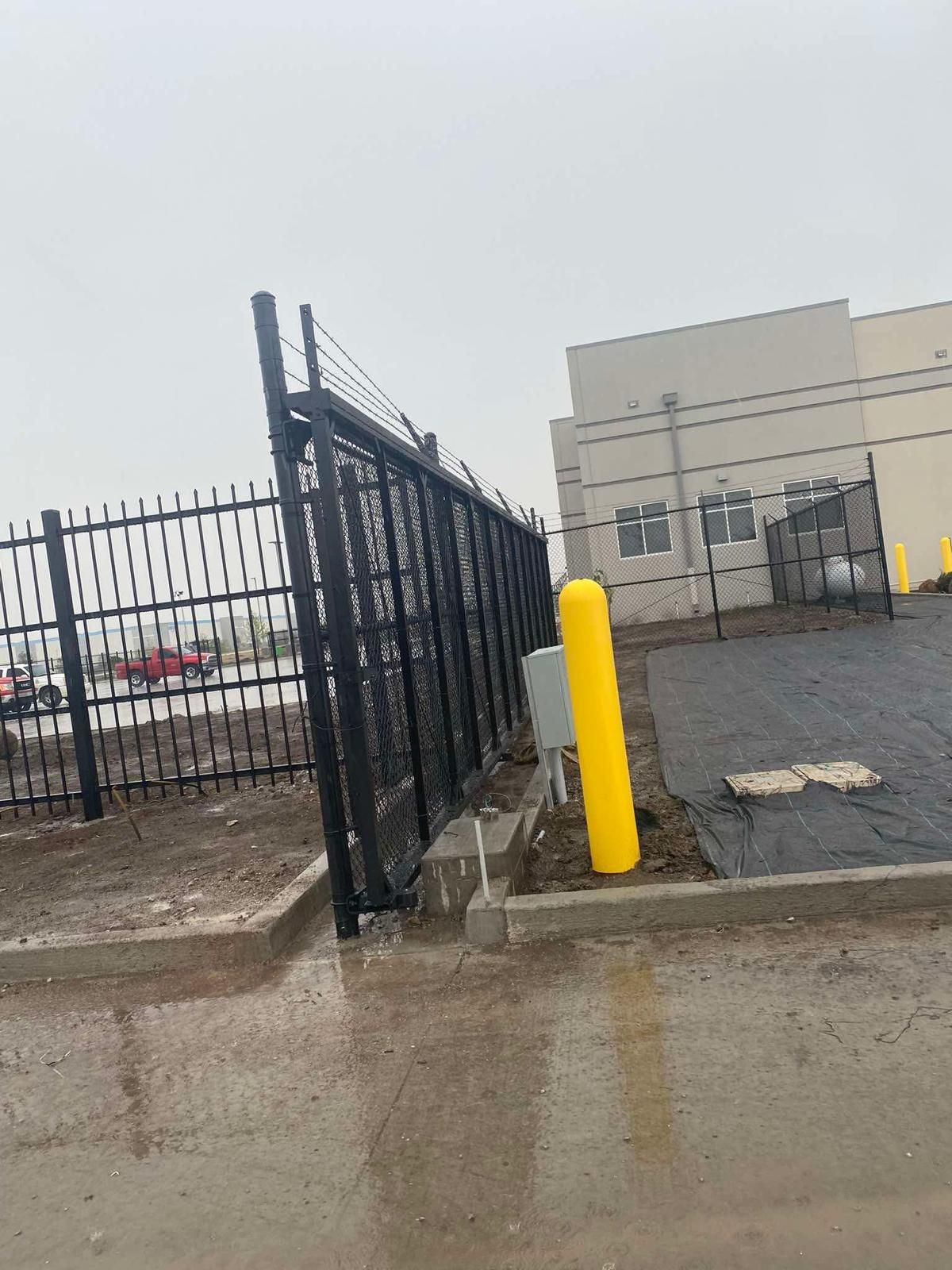 Black metal fence along a concrete curb, with a yellow bollard. A white building and overcast sky are in the background.