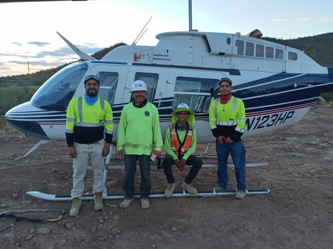 Four workers in safety vests and hard hats stand in front of a white and blue helicopter, possibly for mining work.