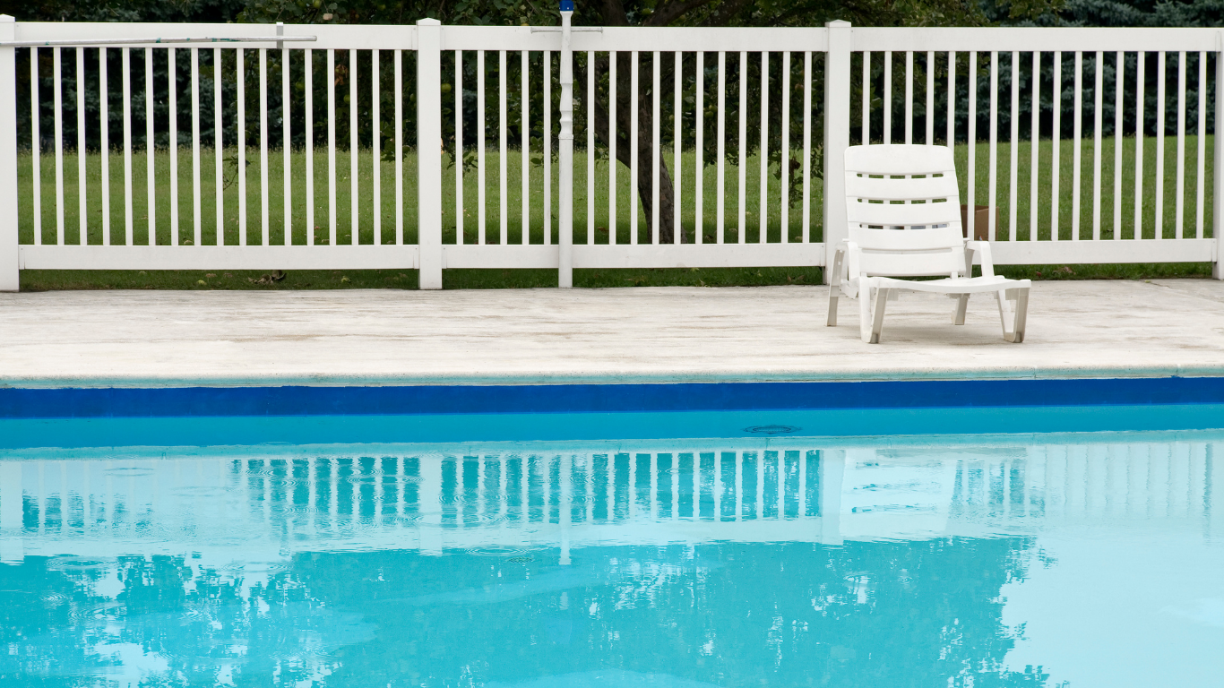 A white fence surrounds a pool, with a lounge chair on the deck. The blue water reflects the sky.