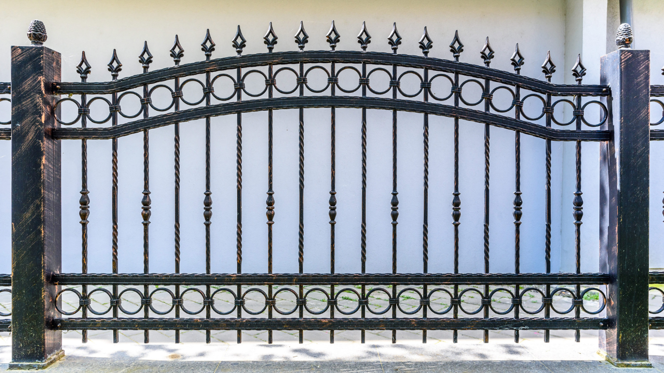 Black wrought iron fence with decorative details between two dark pillars, in front of a white wall.