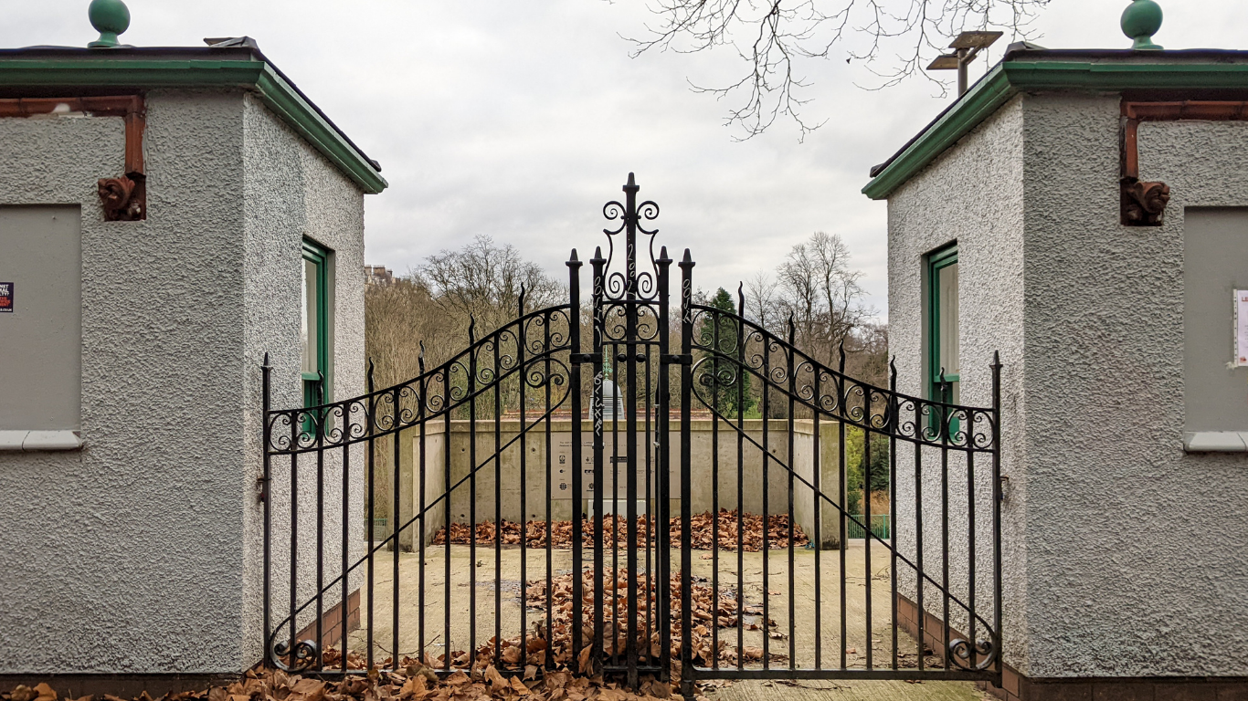 Black wrought-iron gate between two concrete pillars, entrance to a garden or park. Overcast day.
