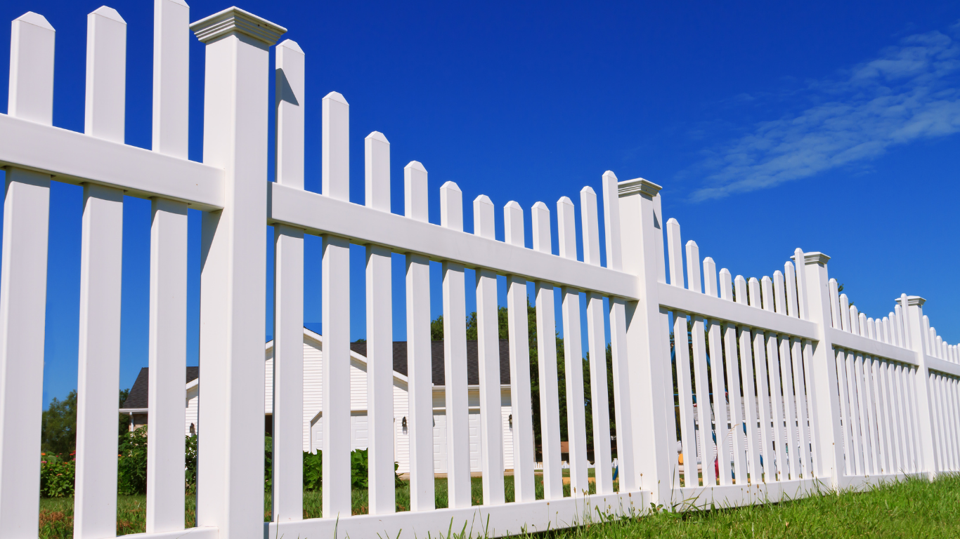 White picket fence against a bright blue sky, with grass in the foreground and a house visible in the distance.