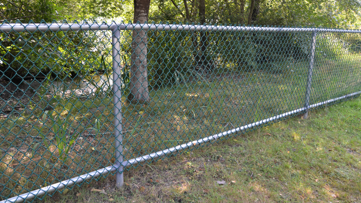 Chain-link fence with green mesh, posts and railings on grass next to trees.