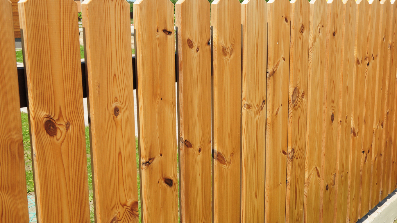 Wooden picket fence, painted with a golden stain, standing outdoors in sunlight.