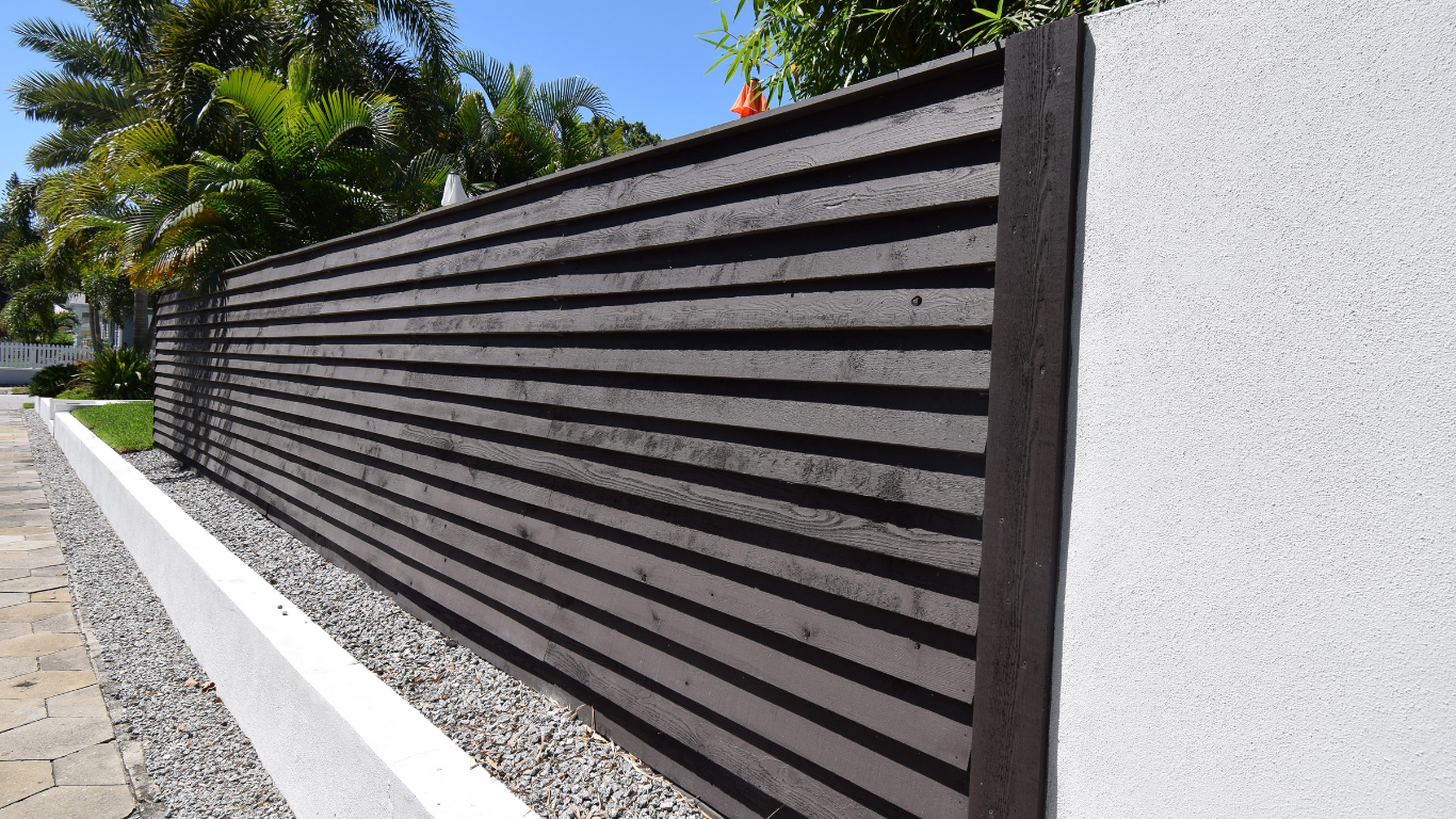 Dark brown horizontal slat fence next to a white wall and a gray gravel border on a sidewalk.