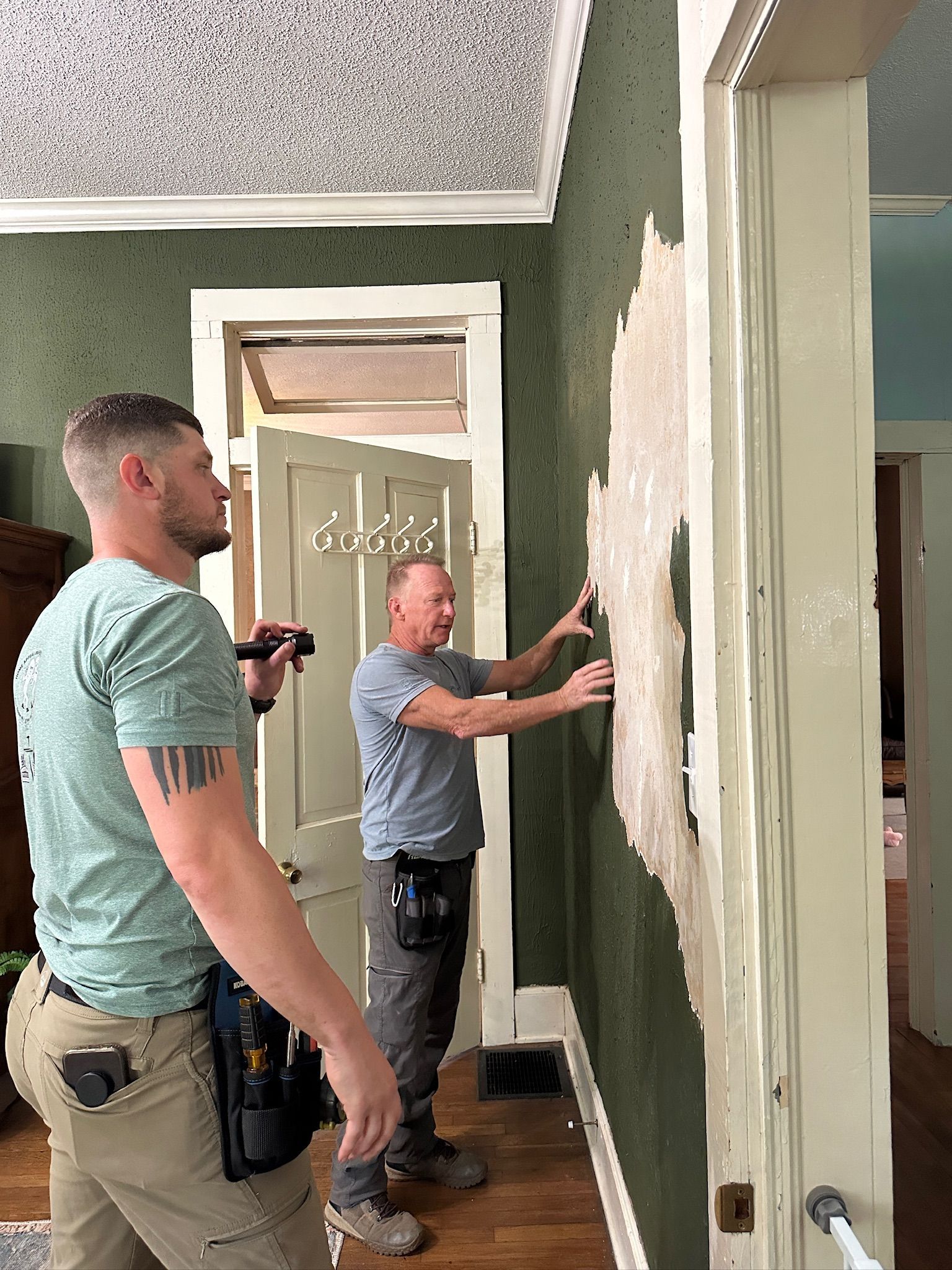 Two men patching peeling wall paint in a green hallway, one holding a trowel.