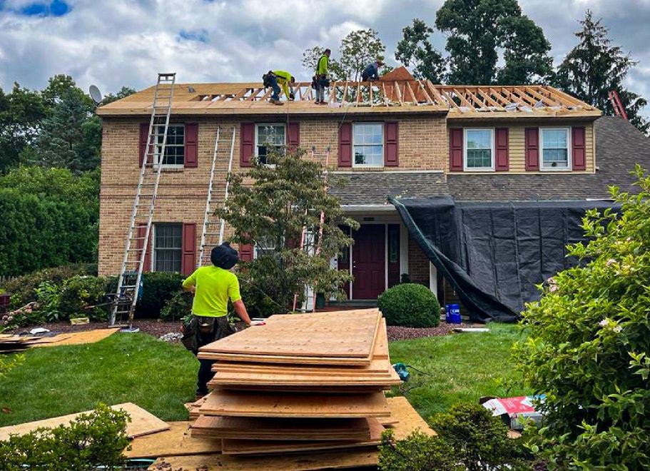 Construction workers replace a roof on a two-story brick house. A worker carries plywood in the foreground, while others work on the roof.