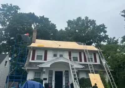 A house undergoing roof repair. Workers use ladders and scaffolding to access the roof, which is partially covered in plywood.
