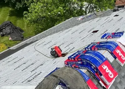 Roofers working on a shingled roof, with equipment and material. Blue and red GAF branded shingle packages are visible, along with a power tool.