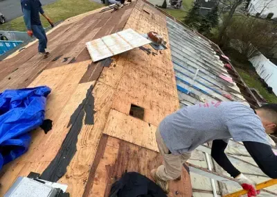 Roofers working on a roof. One is kneeling, another is standing, and the roof is partially stripped.