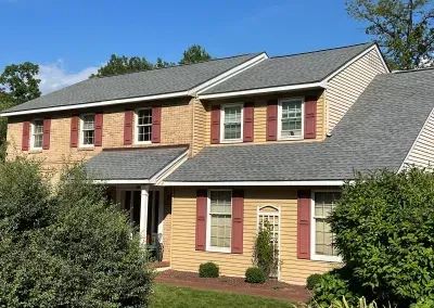 Two-story house with tan siding, a brick front, and a gray shingle roof. Red shutters frame the windows.