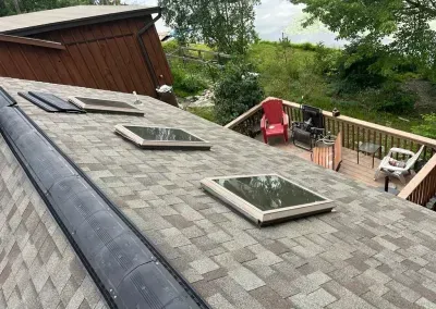 Shingled rooftop with three skylights and a deck in the background. A red chair and trees are visible beyond the deck.