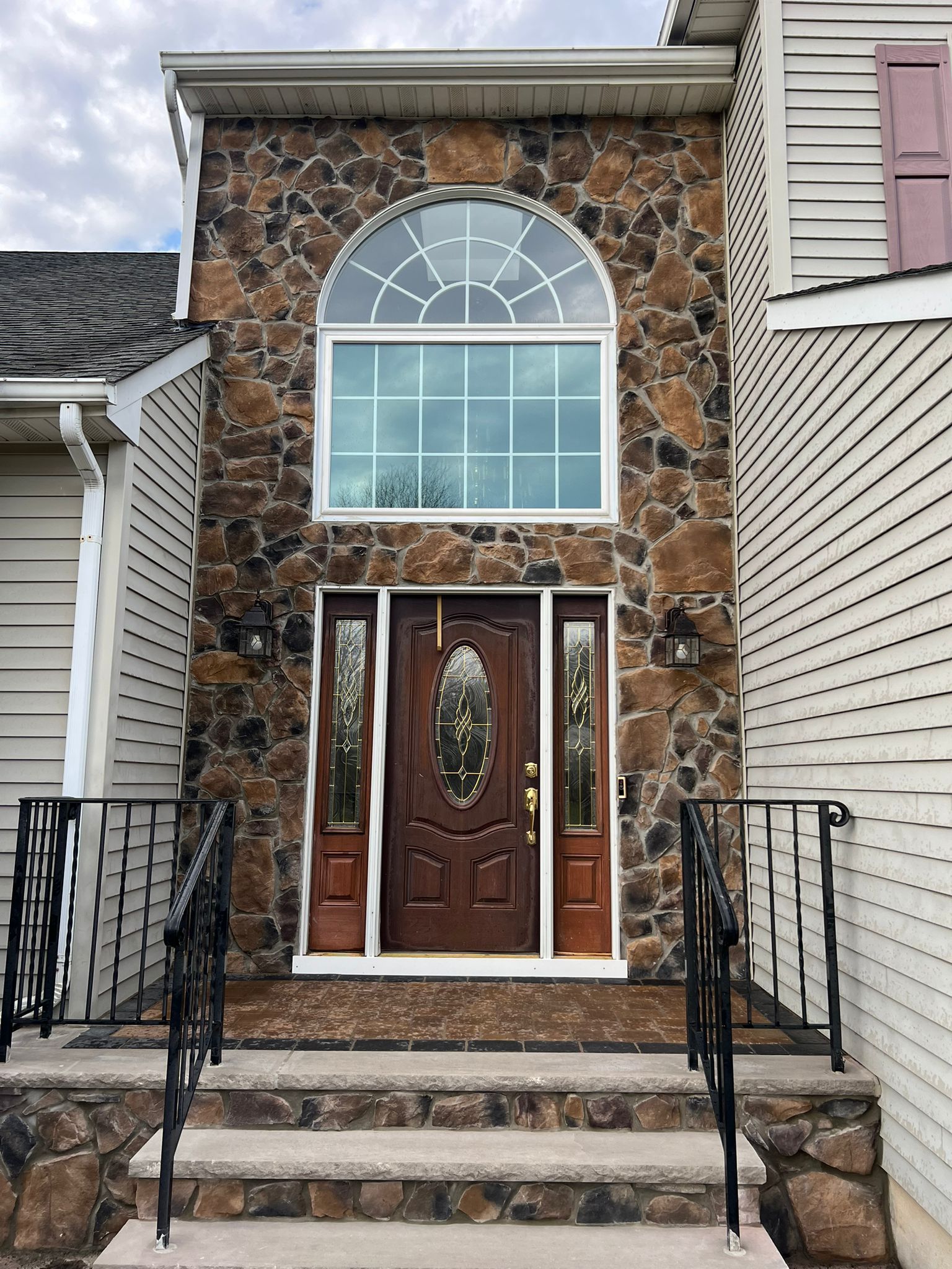 Stone facade entryway with arched window above a brown door and sidelights. Steps lead up to the entrance with black iron railings.